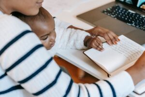 A mother working at a home desk while caring for a young child nearby